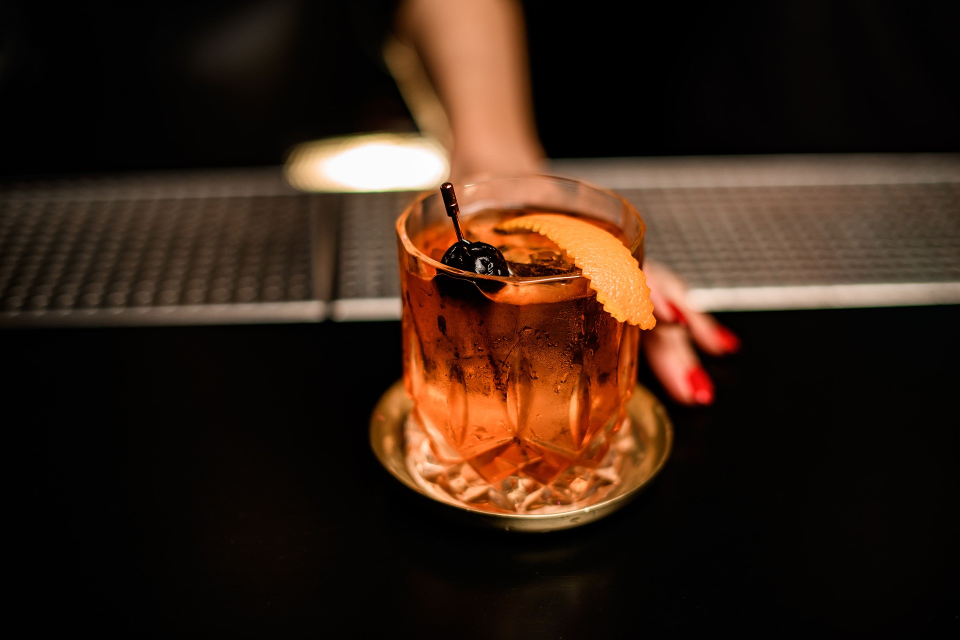 Close-up of old-fashioned glass with alcoholic cocktail to which woman's hand reaches